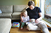 Far og barn sitter sammen i en sofa og leser en illustrert bok i stuen.