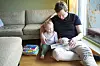 Far og barn sitter sammen i en sofa og leser en illustrert bok i stuen.