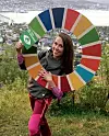 Sarah Lisa Arnesen holder et stort, fargerikt hjul foran et bylandskap og et vannområde.