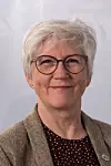 Portrett av en kortklipt kvinne med briller, brun jakke og mørk bluse mot lys bakgrunn.