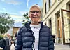 Smilende eldre kvine med kort hår, briller og blå dunjakke står i en bygate.