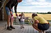 Et lite barn står mellom to voksne på en treveranda ved et gårdstun.