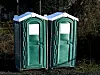 To grønne mobile toaletter står utendørs foran et skogsområde.