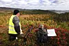 Jeger i synlighetsvest sitter i lyngen med våpen og sekk foran et høstfarget fjellandskap.
