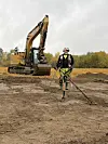 En gravemaskin i bakgrunnen på et jorde. En metalldetektorist står midt i bildet og søker med metalldetektor i jorda.