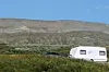 En hvit campingvogn og en mørk bil står foran en lav, vegetasjonskledd fjellrygg.