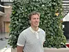 Smilende mann i stripet skjorte står foran en vegg med grønne planter.
