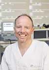 Smilende mann i hvit medisinsk drakt foran skjermer og medisinsk utstyr.