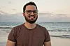 En smilende mann med briller står foran havet på en strand.