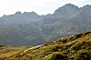 Grønn fjellside i forgrunnen med bratte, lagdelte fjell og en høy fjellkjede i bakgrunnen.