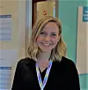 Smilende blond kvinne, Tonje Amland i svart genser med fargerik navnesnor foran en glassdør.