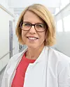Smilende kvinne med briller og hvit frakk i en lys korridor.