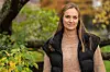 Kvinne i svart vest og strikket genser står utendørs blant grønne og høstfargede planter.