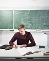 Lærer skriver i en bok ved et skrivebord foran en grønn tavle.