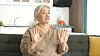 Eldre kvinne sitter i en mørk sofa og gestikulerer med begge hendene.