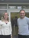 To smilende voksne står foran glassdører ved et moderne bygg.