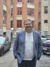 Smilende mann i blå blazer foran teglsteinsbygninger og parkerte biler.