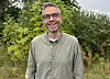 Smilende mann med briller og grågrønn skjorte foran grønne busker.