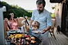 Far og sønn griller sammen på en terrasse, med grillmat i forgrunnen og familie i bakgrunnen.