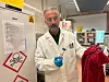 Awat Dostberg holder en rød kjole i fløyel i den ene hånda, og et reagensrør i den andre hånda. Han står inne på laboratoriet sitt foran en dør med fare for gift-skilt foran. Han har på seg hvit laboratoriefrakk, laboratoriebriller og blå gummihandsker.