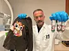 Awat Dostberg holder opp en svart genser med en ugle i paljetter i den ene hånda, og tre reagensrør i den andre hånda. Han er på et laboratorie og har på seg hvit labfrakk og laboratoriebriller.