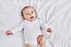 En søt baby i hvit body ligger på et hvitt laken og smiler i kameraet.