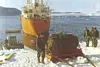 Den store meteoritten står innpakket i et nett på en slede over snøen nær vannet. Tre menn står på den ene siden og en på den andre. Et stort, oransje skip ligger fortøyd i bakgrunnen.