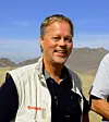 Portrettbilde av Axel Christophersen. Han har svart skjorte og en beige vest. Det henger solbriller rundt halsen hans, og i bakgrunnen kan vi se blå himmel og fjell. Han er i Afghanistan.