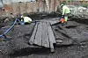 Midt i bildet ligger fire store treplanker som danner en vei fra middelalderen. Bakerst på bildet på hver side av veien står to arkeologer. Plankeveien fortsetter ut av bildet til høyre.