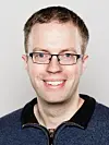 Smilende mann med briller og kort mørkt hår fotografert mot lys bakgrunn.