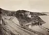 Historisk svart-hvitt bilde av jernbanespor, hus og vann langs et kupert landskap.