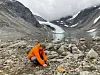 Mann i oransje jakke sitter på huk blant steiner ved et turkis brevann omgitt av fjell.