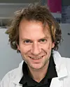 Smilende mann med krøllete hår iført hvit frakk foran en lys innendørs bakgrunn.