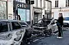 Utbrente biler og vrakrester i en Paris-gate, ved butikkfasader, med en kvinne som står og ser på med måpende ansiktsuttrykk.