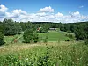 Frodig eng og gård omgitt av skog under en blå himmel med hvite skyer.
