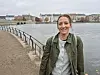 Bilde av Anne Lovise Nordstoga som står ved en vannkant med bybebyggelse og rekkverk i bakgrunnen.