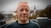 Portrett av en mann i mørk genser, fotografert utendørs med bybebyggelse og trær i bakgrunnen.