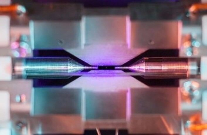 Her kan du skimte den ørlille prikken av et strontium-atom der det ligger badet i blå-fiolett laserlys og i spenn i det elektriske feltet mellom de to elektrodene. Strontium-atomer som dette blir brukt i supernøyaktige atomklokker. (Foto: David Nadlinger ved University of Oxford) Her kan du skimte den ørlille prikken av et strontium-atom der det ligger badet i blå-fiolett laserlys og i spenn i det elektriske feltet mellom de to elektrodene. Strontium-atomer som dette blir brukt i supernøyaktige atomklokker. (Foto: David Nadlinger ved University of Oxford)